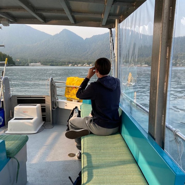 Person seated on a boat looking at distant mountains through binoculars.