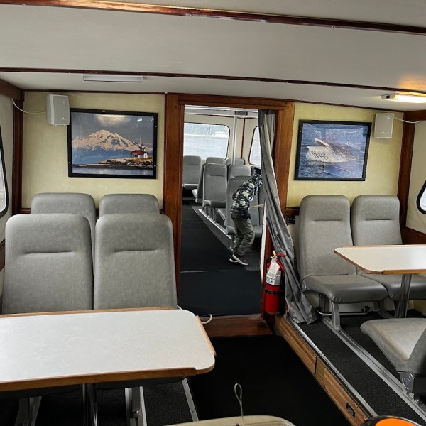 Interior of a boat with seating, two tables, framed pictures, and a fire extinguisher.