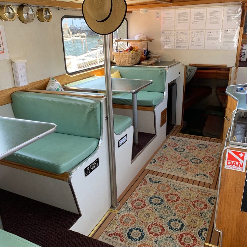 Boat interior with mint green seating, a straw hat hanging, and colorful patterned rugs on wood floor.